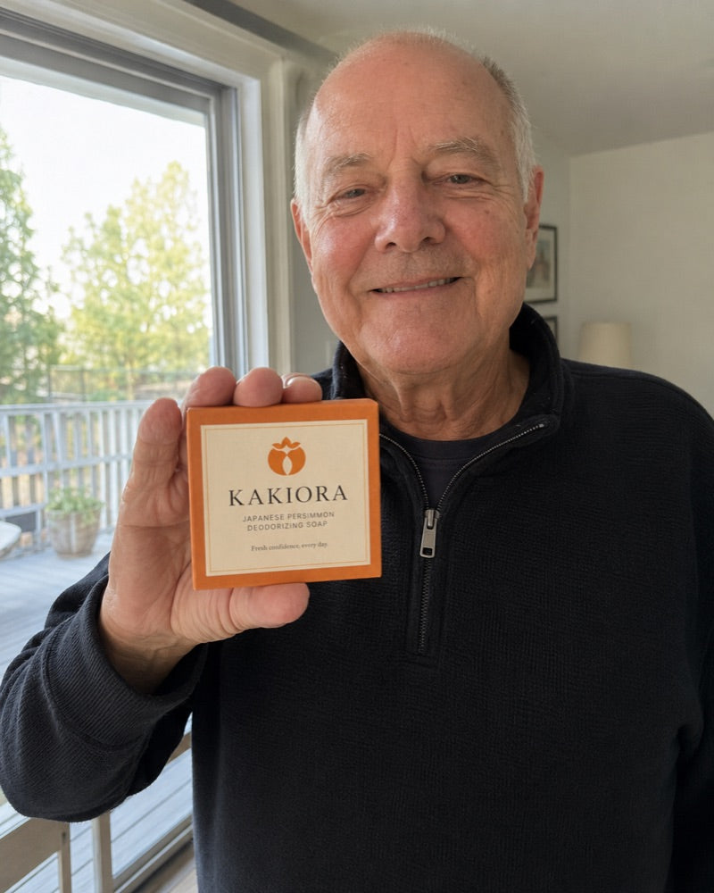 Older man holding a box of KAKIORA persimmon soap by a bright window